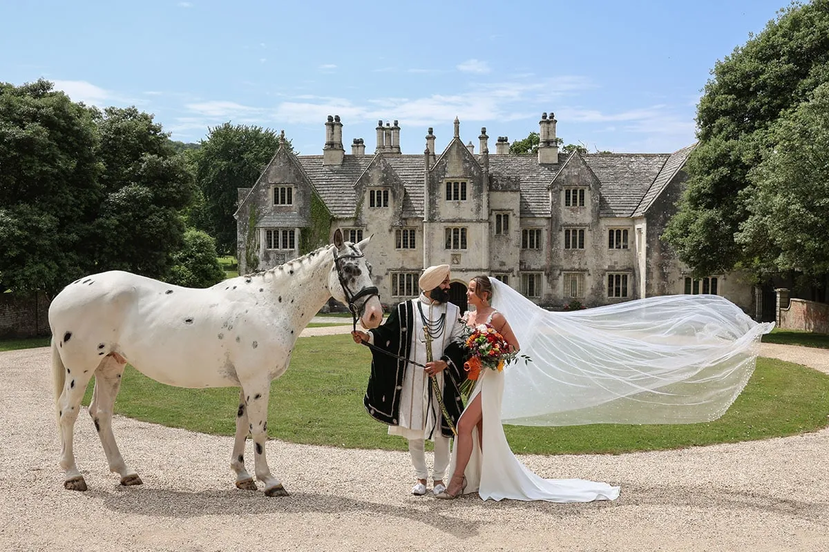 Bride's veil catching the wind beside a white horse at a manor house